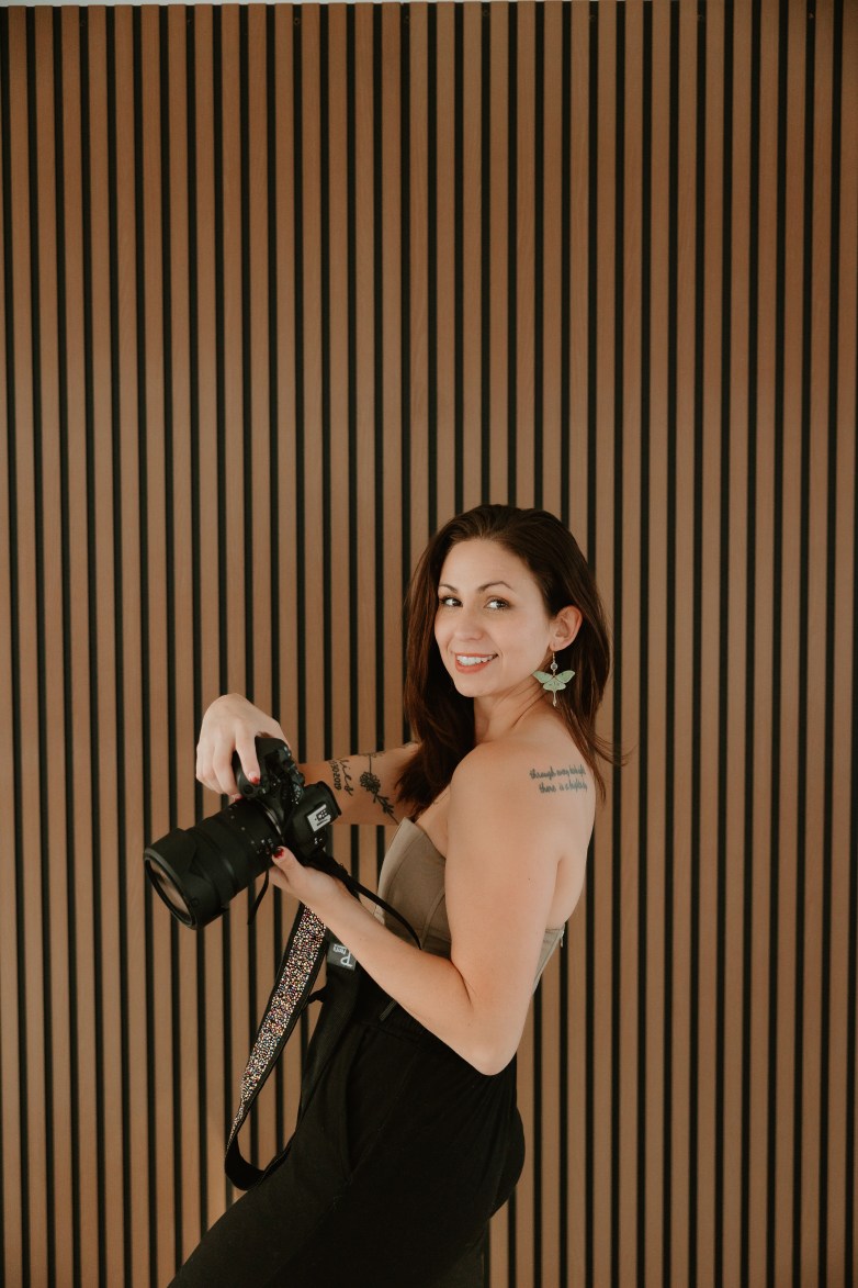 ARTographer Klohe in a green strapless corset top holding a Canon camera against a brown wall.